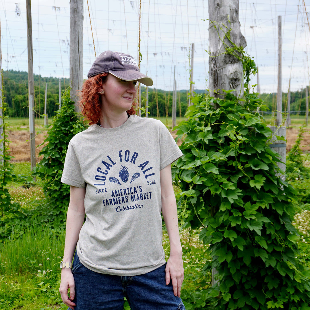 A woman with red hair wearing a light gray graphic t-shirt and a baseball cap stands in a lush green farm field with trellis and climbing plants in the background. The t-shirt features a navy blue graphic that reads 'LOCAL FOR ALL' and 'AMERICA'S FARMING MARKET Collaboration'.