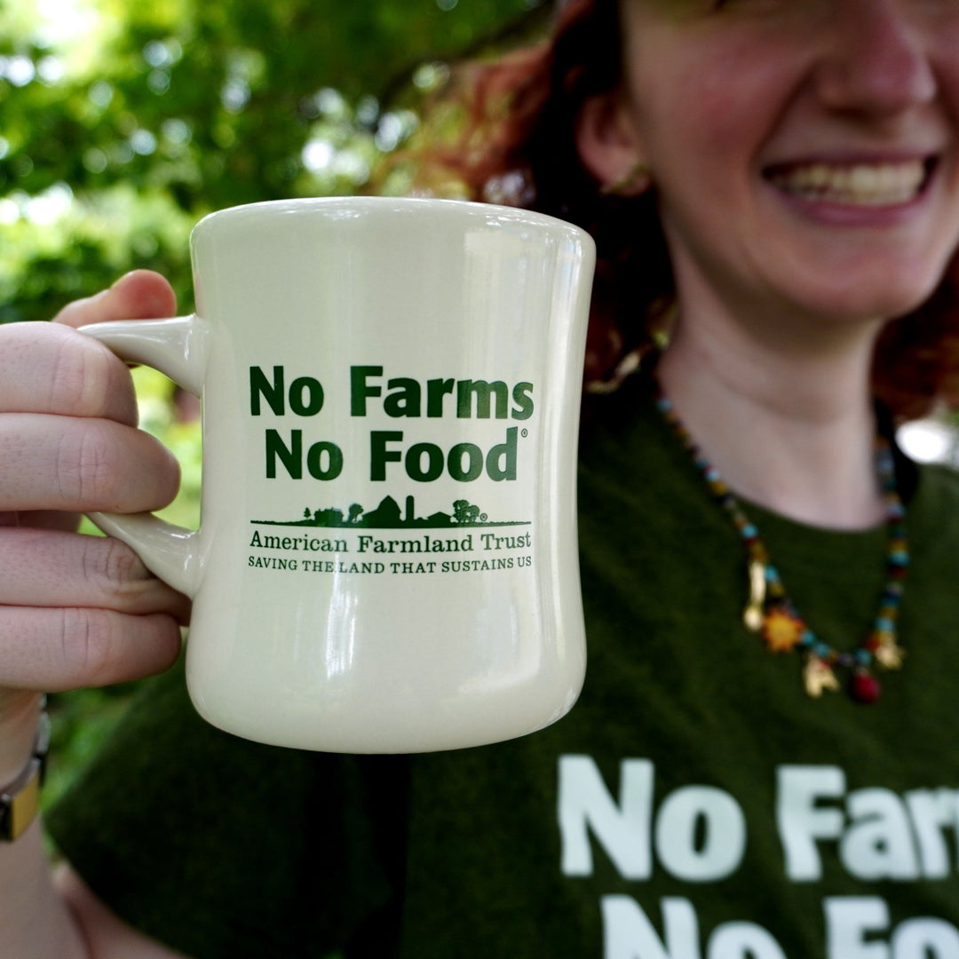 A person outdoors holds up a cream-colored ceramic mug with the text 'No Farms No Food' and 'American Farmland Trust' printed in green. The person is wearing a matching dark green t-shirt with the same slogan.