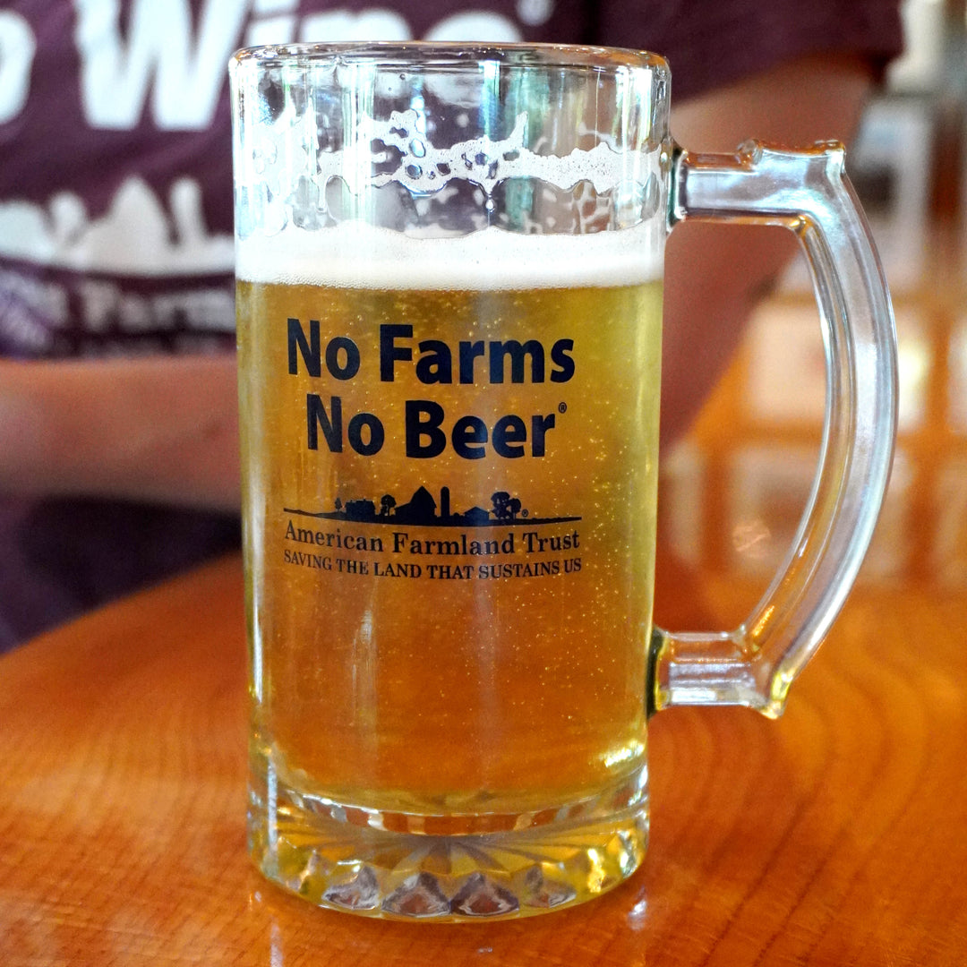A clear glass beer mug filled with golden beer and topped with white foam, sitting on a wooden table. The mug has a black printed logo that reads 'No Farms No Beer' with the American Farmland Trust underneath.